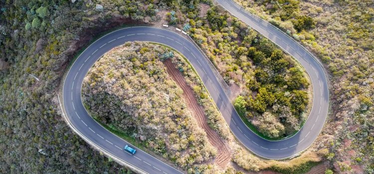 An aerial shot of a car passing through a spiral road surrounded by trees in the countryside