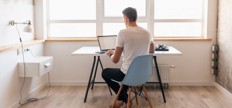 young handsome man in casual outfit sitting at table working on laptop, freelancer at home, view from back